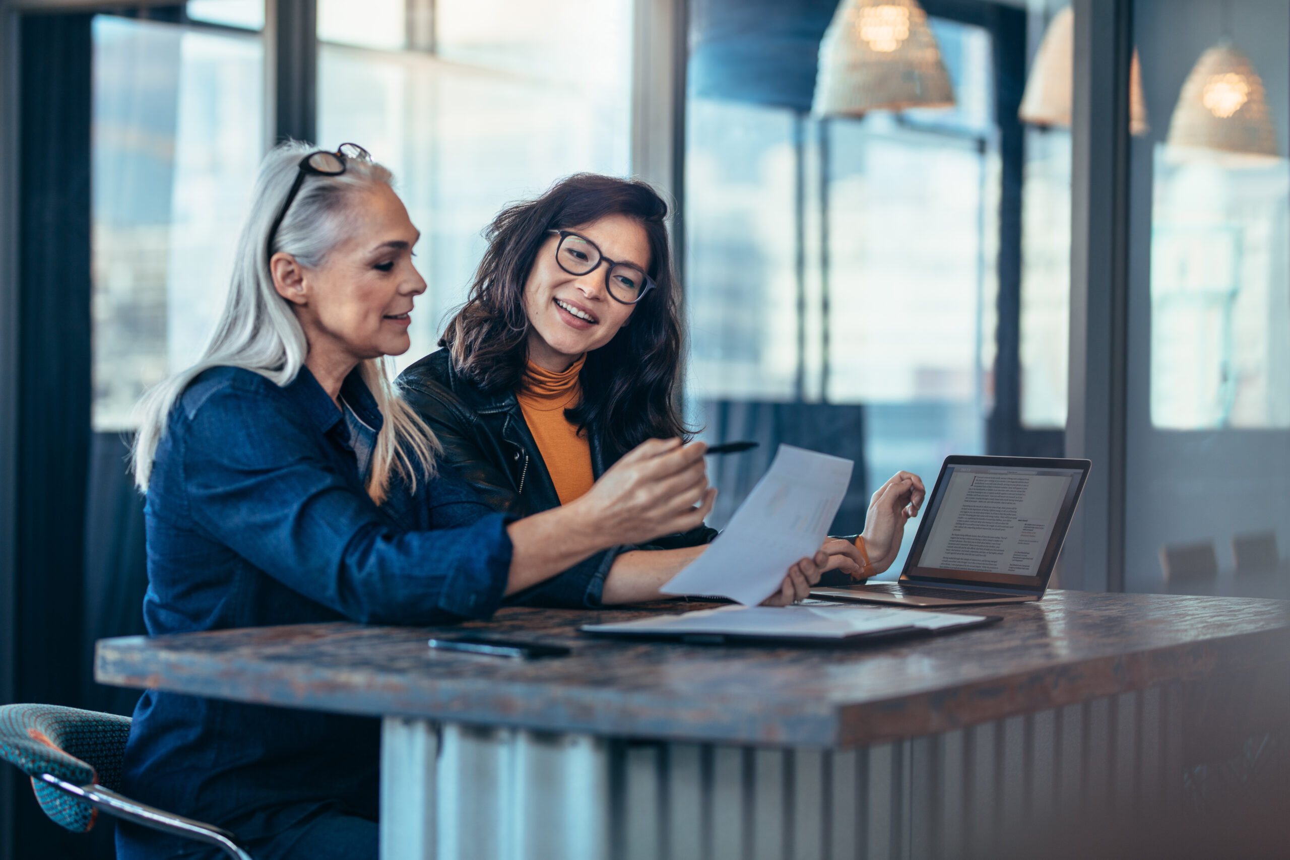 Two,Women,Analyzing,Documents,While,Sitting,On,A,Table,In